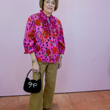 Woman wearing a colorful patterned blouse and holding a black handbag with a white bow against a pink wall.