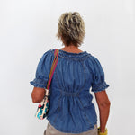An older woman viewed from behind wearing a blue denim short-sleeve blouse with puff sleeves and a gathered back, paired with khaki pants and carrying a colorful patterned shoulder bag against a plain white background.