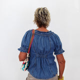 An older woman viewed from behind wearing a blue denim short-sleeve blouse with puff sleeves and a gathered back, paired with khaki pants and carrying a colorful patterned shoulder bag against a plain white background.