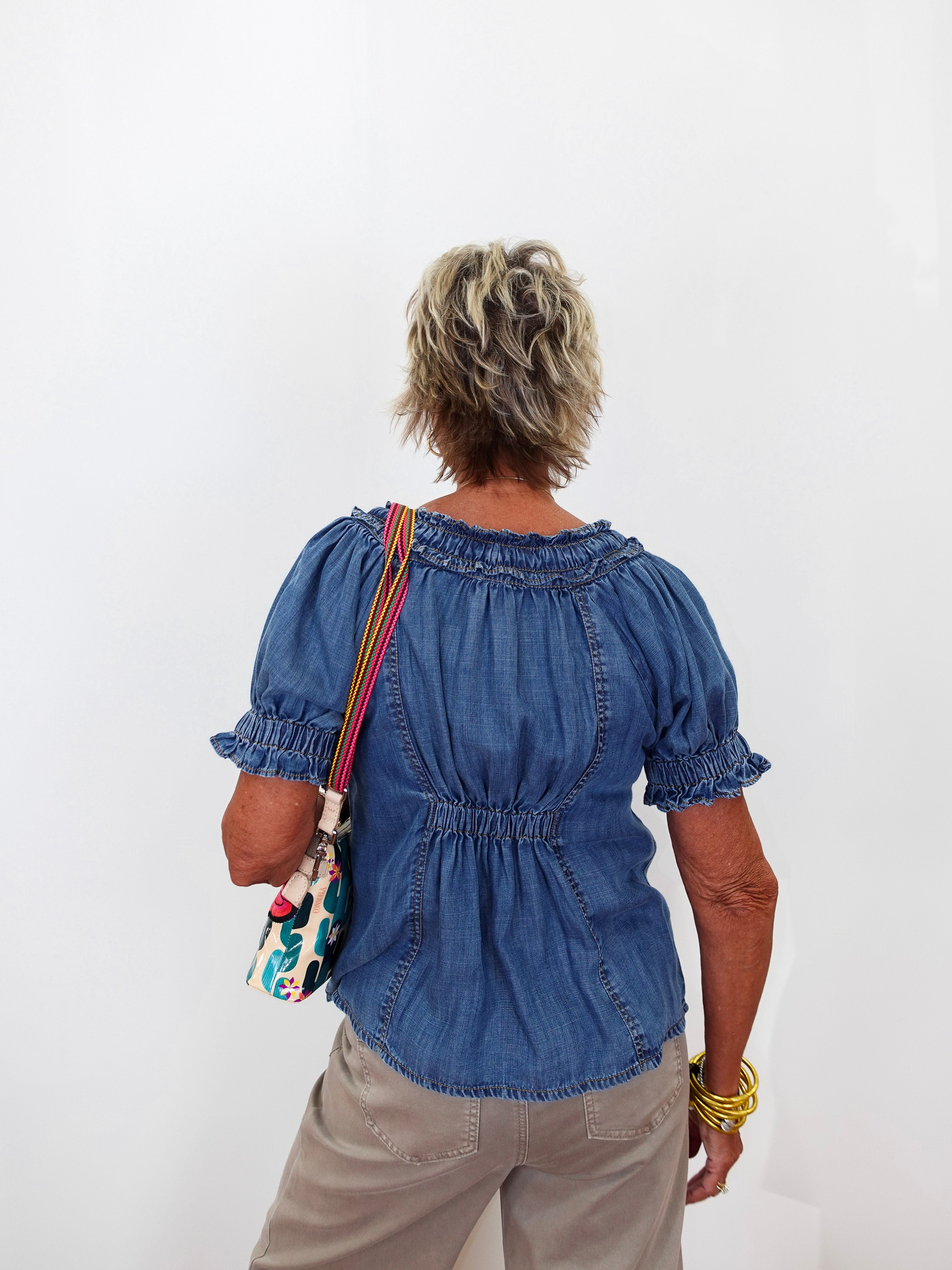 An older woman viewed from behind wearing a blue denim short-sleeve blouse with puff sleeves and a gathered back, paired with khaki pants and carrying a colorful patterned shoulder bag against a plain white background.