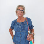 A woman with short gray hair and glasses wearing a blue denim short-sleeve top and beige pants, holding a colorful crossbody bag against a plain white background.