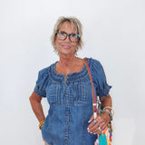 A woman with short gray hair and glasses wearing a blue denim short-sleeve top and beige pants, holding a colorful crossbody bag against a plain white background.