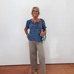A woman stands against a plain white wall wearing a blue denim short-sleeve blouse with puff sleeves, beige cropped trousers, and beige loafers. She is holding a small colorful crossbody bag.