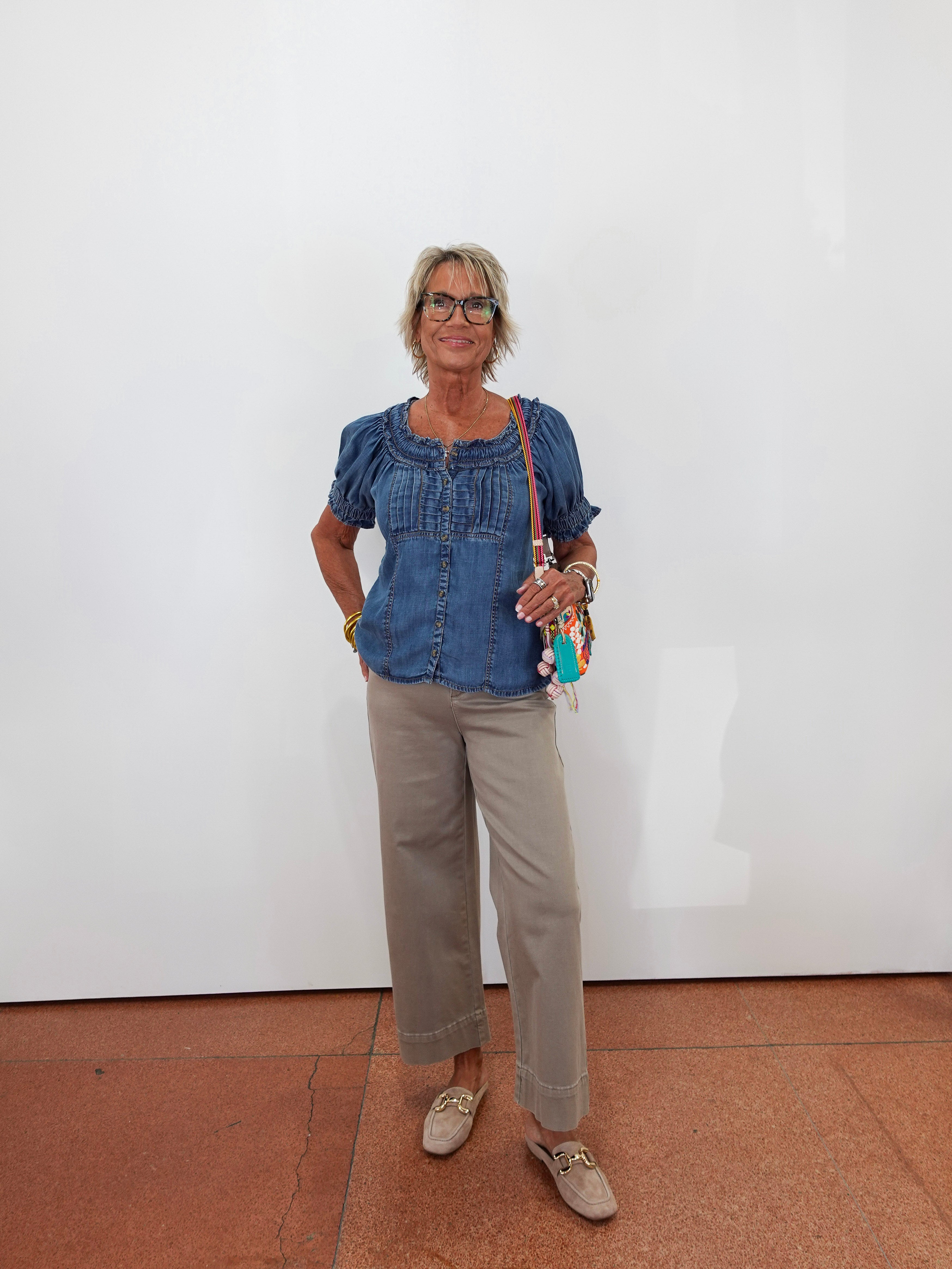 A woman stands against a plain white wall wearing a blue denim short-sleeve blouse with puff sleeves, beige cropped trousers, and beige loafers. She is holding a small colorful crossbody bag.