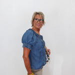 A woman with short blonde hair and glasses, wearing a blue denim puff-sleeve top and beige skirt, holding a small purse against a plain white background.