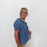 A woman with short blonde hair and glasses, wearing a blue denim puff-sleeve top and beige skirt, holding a small purse against a plain white background.
