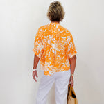 Person wearing an orange and white patterned shirt with white pants, standing against a white wall.