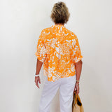 Person wearing an orange and white patterned shirt with white pants, standing against a white wall.