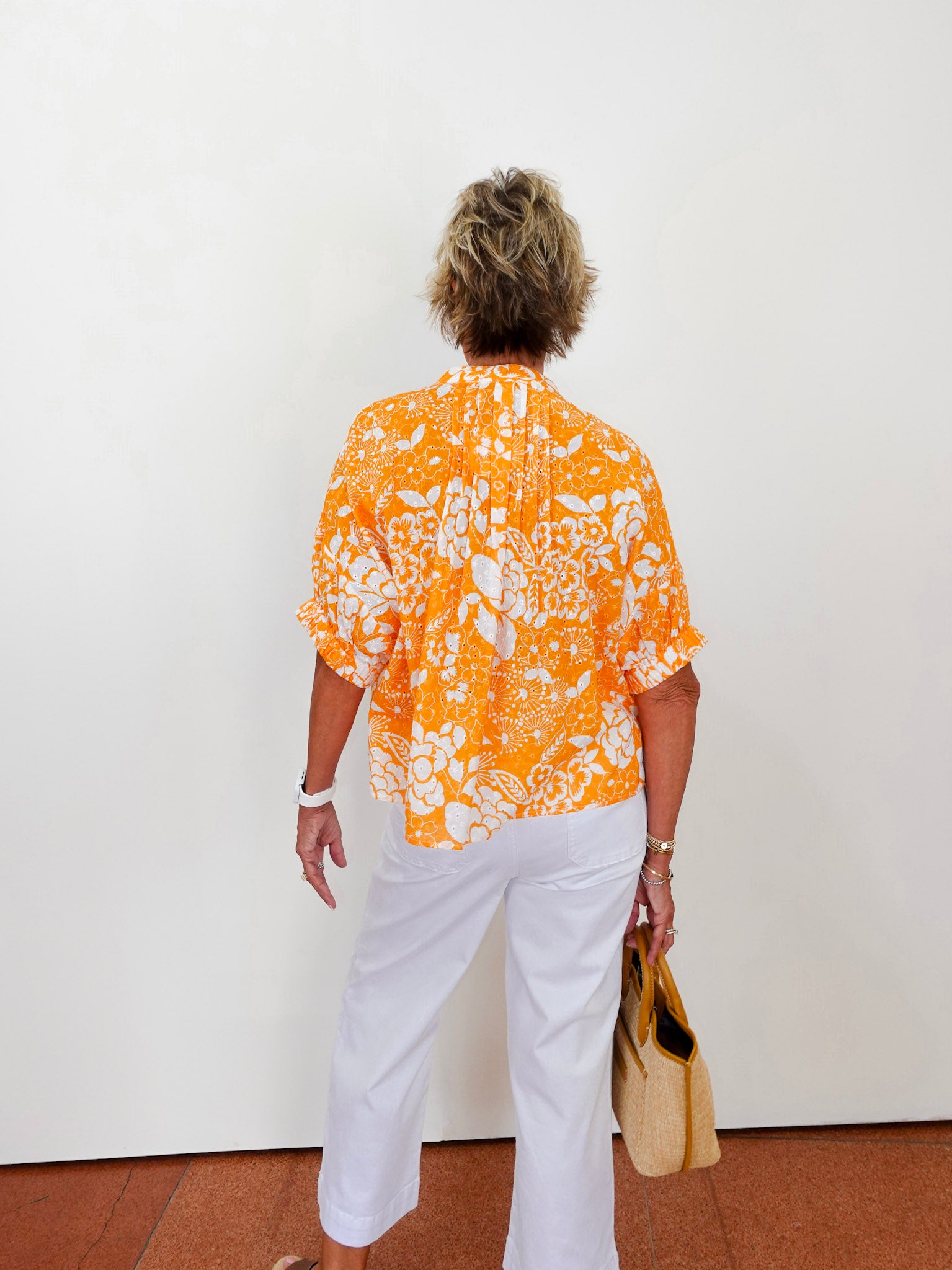 Person wearing an orange and white patterned shirt with white pants, standing against a white wall.