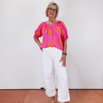 Woman wearing a bright pink blouse with orange patterns and white pants against a white wall.