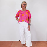 Woman wearing a bright pink blouse with orange patterns and white pants against a white wall.