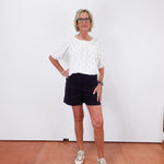 Woman wearing a white blouse and black shorts standing against a white wall.