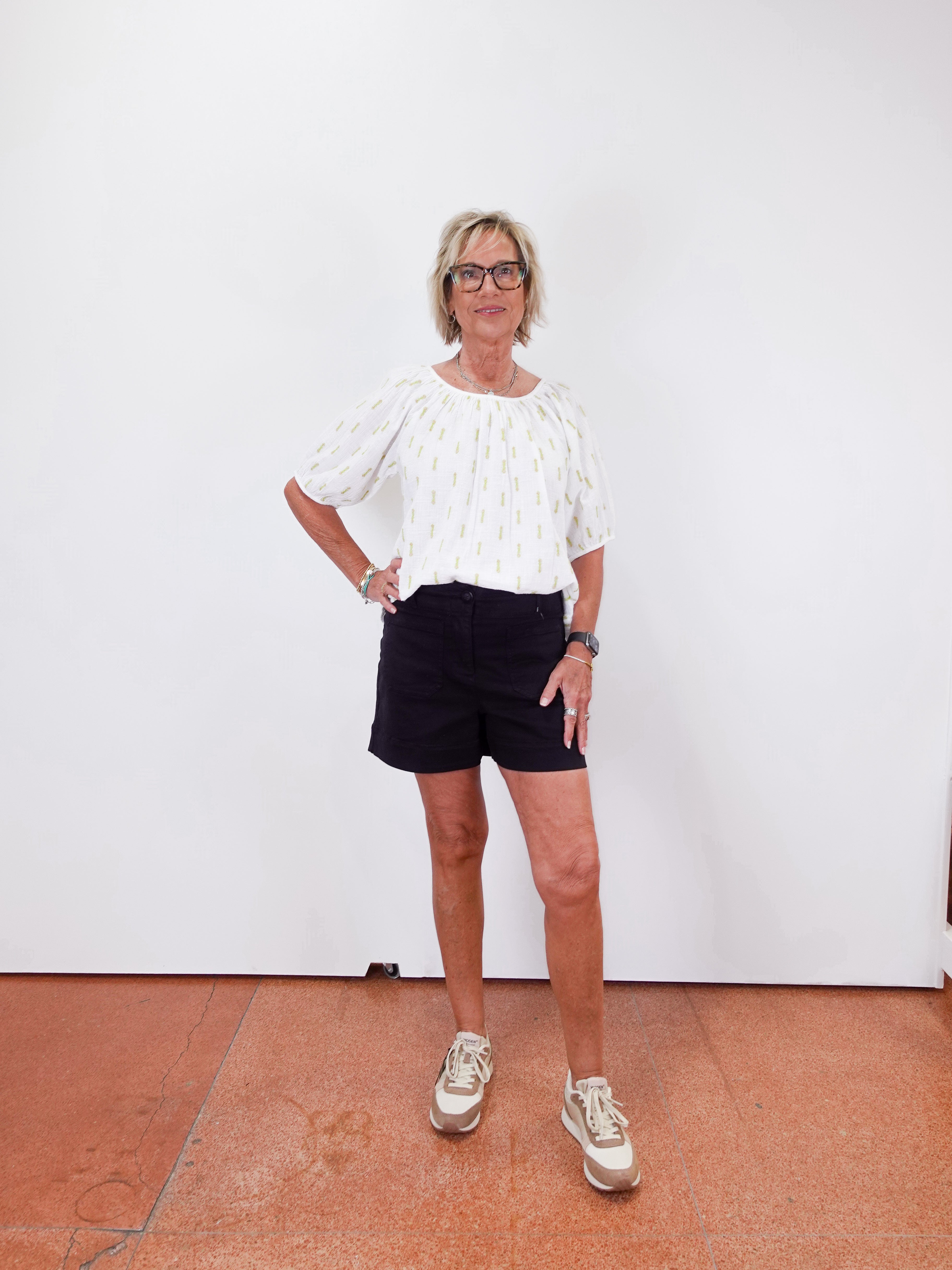 Woman wearing a white blouse and black shorts standing against a white wall.
