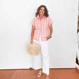Woman wearing a striped shirt and white pants, holding a hat indoors.