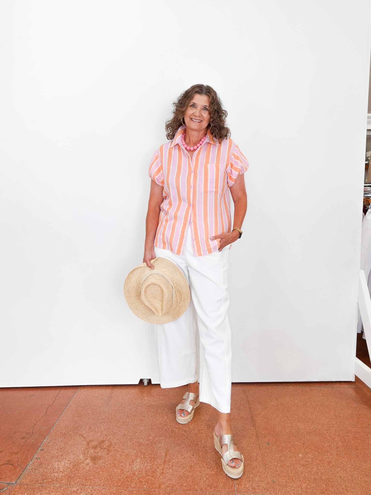 Woman wearing a striped shirt and white pants, holding a hat indoors.