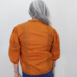 A person viewed from behind wearing a burnt orange blouse with embroidered details on the back and sleeves, paired with blue jeans, against a plain white wall.