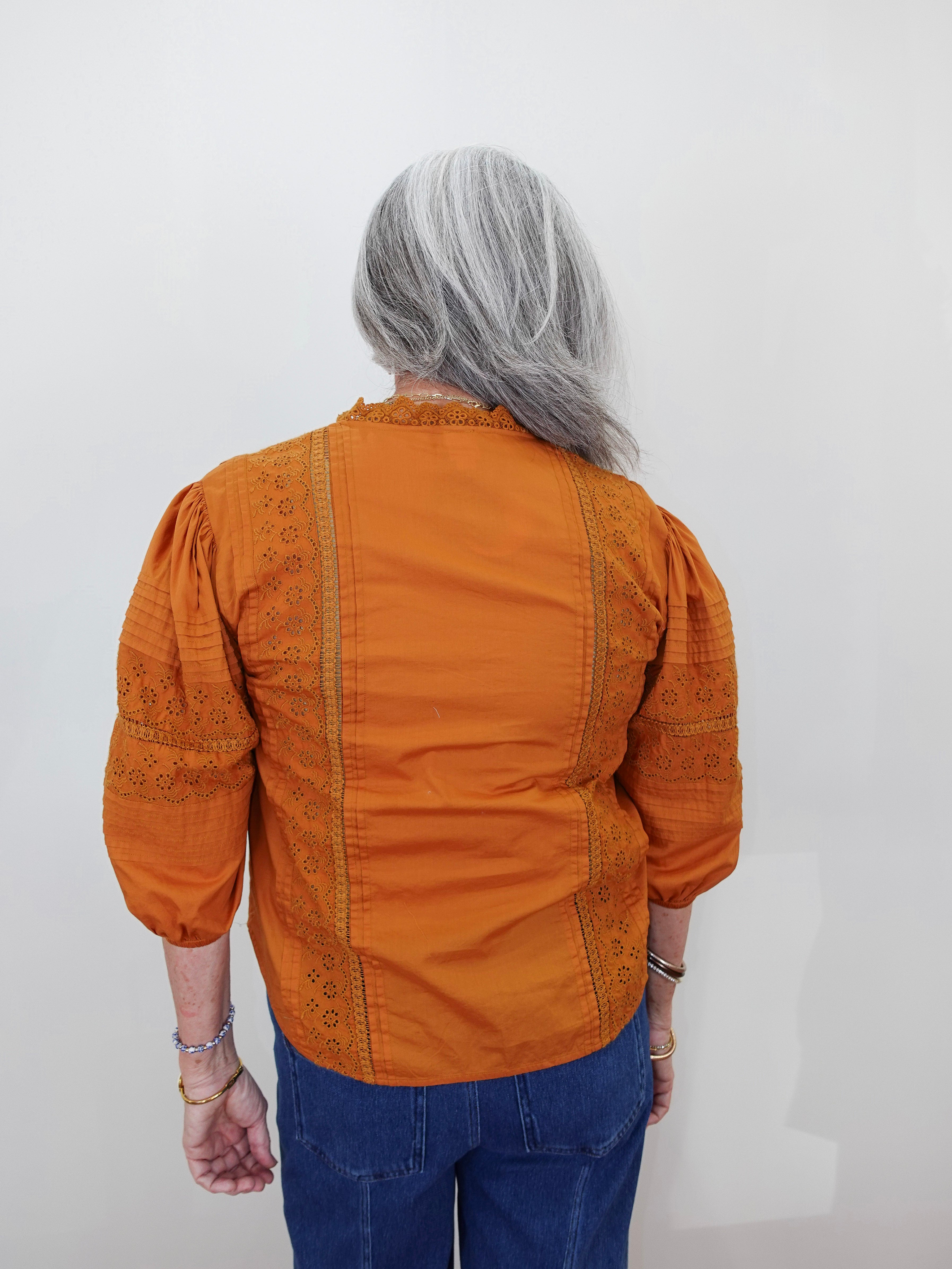 A person viewed from behind wearing a burnt orange blouse with embroidered details on the back and sleeves, paired with blue jeans, against a plain white wall.