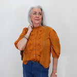 An older woman with gray hair is wearing a textured mustard orange blouse with ruffle details and blue jeans, standing against a plain white wall.