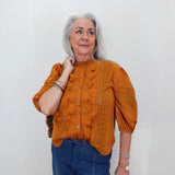 An older woman with gray hair is wearing a textured mustard orange blouse with ruffle details and blue jeans, standing against a plain white wall.