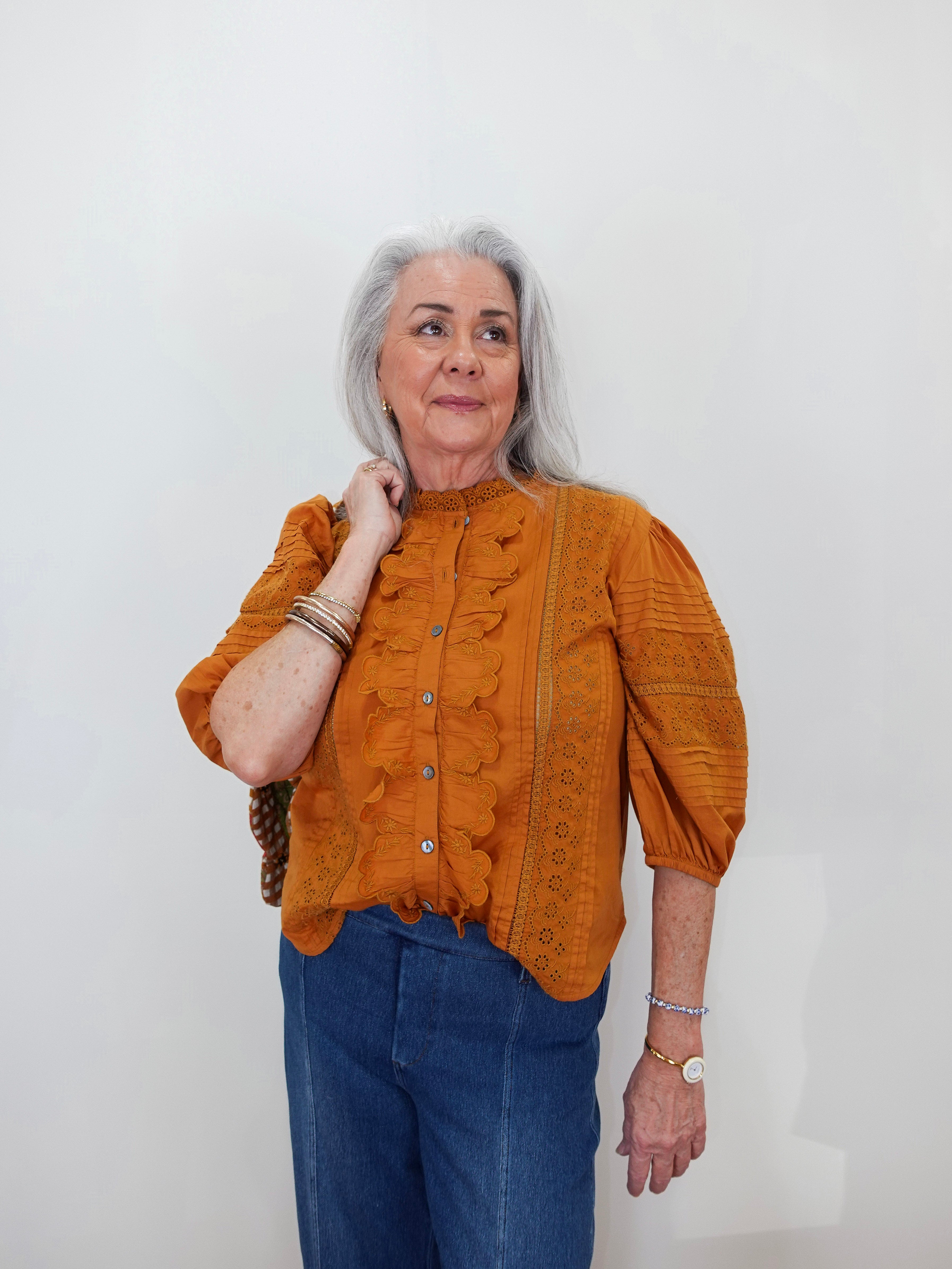 An older woman with gray hair is wearing a textured mustard orange blouse with ruffle details and blue jeans, standing against a plain white wall.