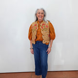An older woman with gray hair stands against a plain white wall wearing a rust-orange blouse, a white vest with orange and red floral embroidery, blue cropped jeans, and beige platform sandals.