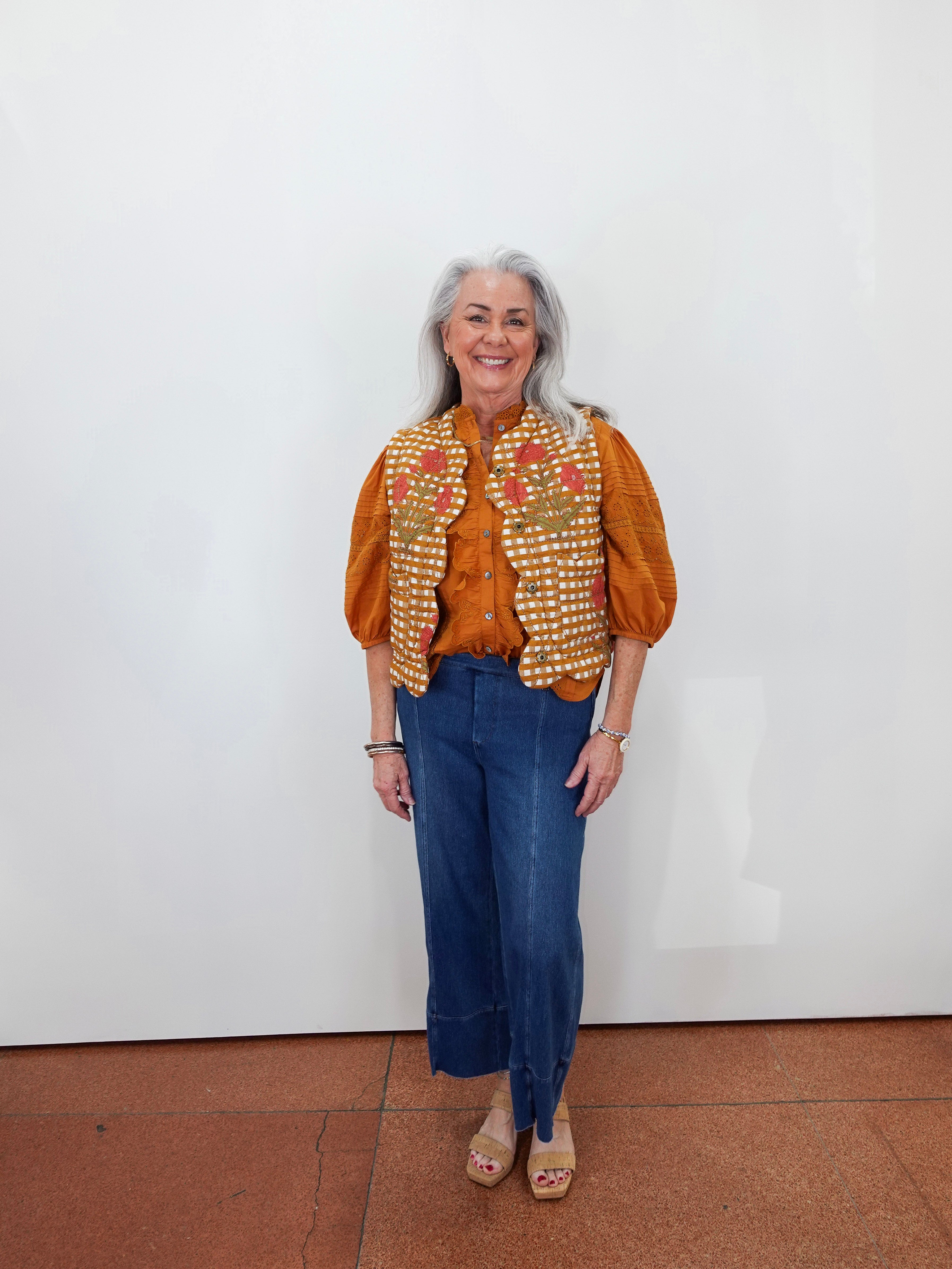 An older woman with gray hair stands against a plain white wall wearing a rust-orange blouse, a white vest with orange and red floral embroidery, blue cropped jeans, and beige platform sandals.