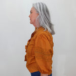 A woman with shoulder-length gray hair is shown in profile wearing a rust-orange embroidered blouse and blue jeans against a plain off-white background.