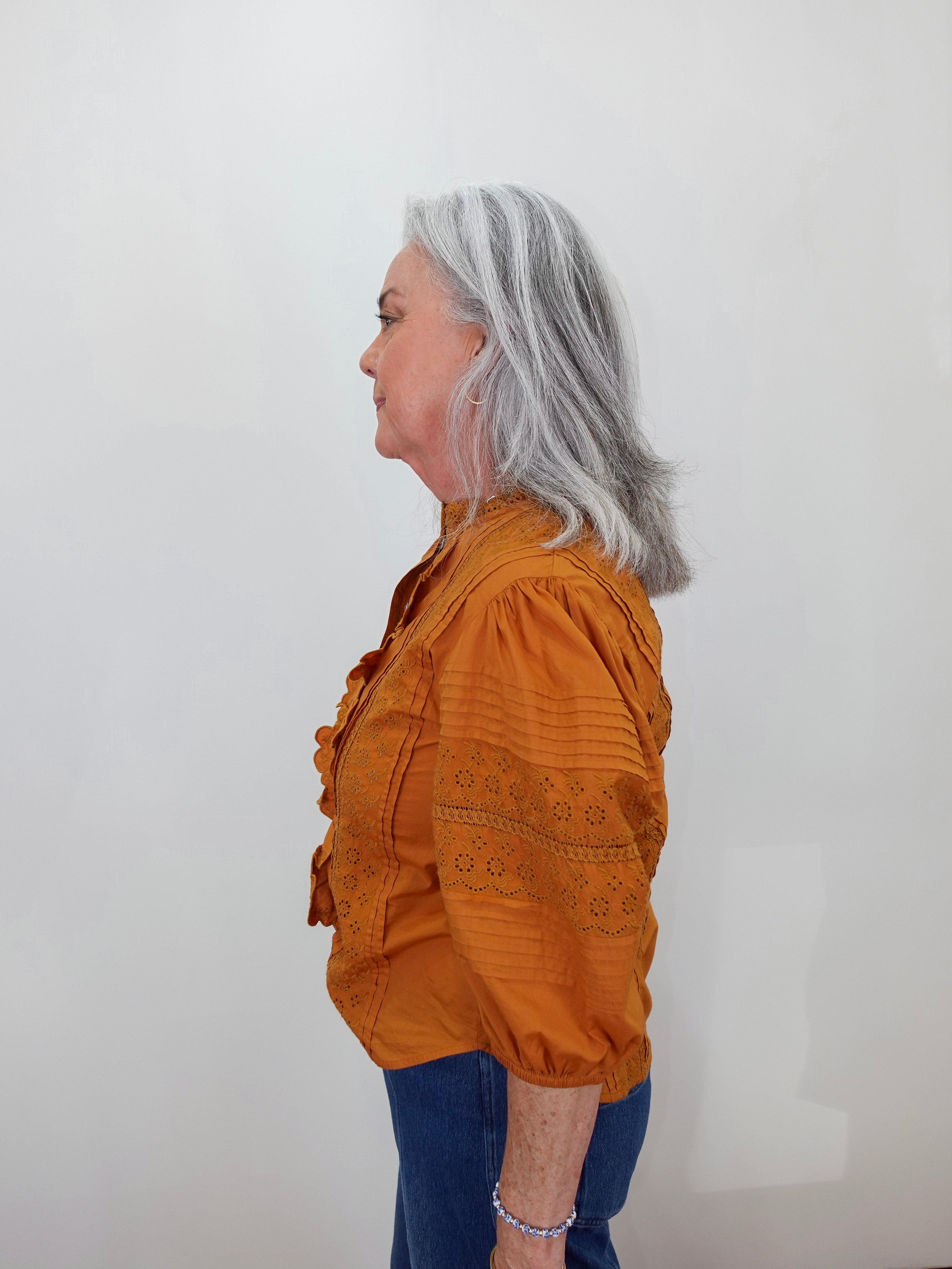 A woman with shoulder-length gray hair is shown in profile wearing a rust-orange embroidered blouse and blue jeans against a plain off-white background.