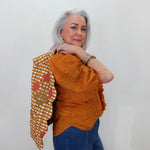 An older woman with gray hair is shown from the waist up, wearing a rust-orange embroidered blouse and blue jeans, with a patterned orange and white checkered shawl draped over her left shoulder. She is standing against a plain white wall.