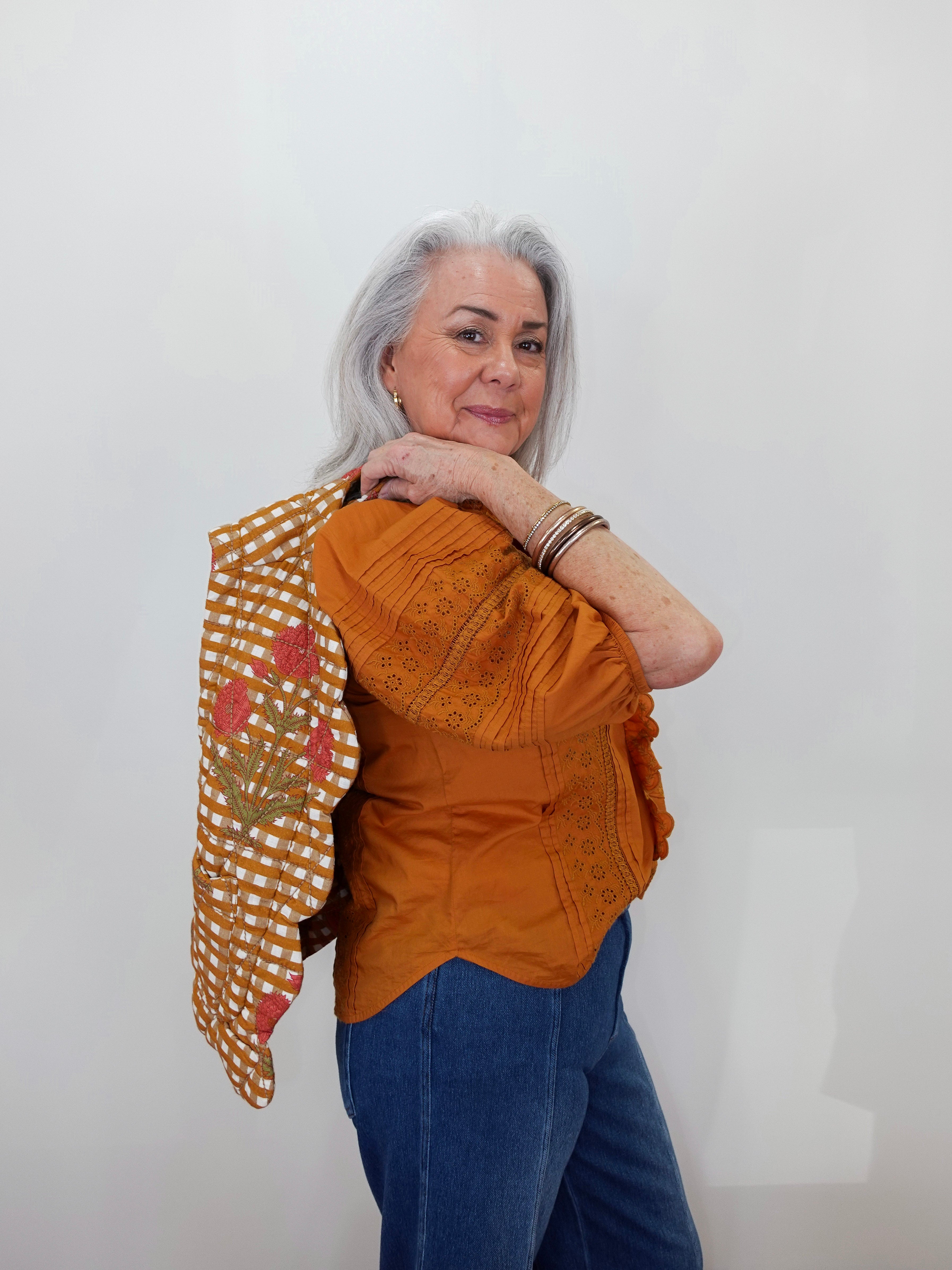 An older woman with gray hair is shown from the waist up, wearing a rust-orange embroidered blouse and blue jeans, with a patterned orange and white checkered shawl draped over her left shoulder. She is standing against a plain white wall.