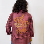 Person wearing a red plaid shirt with gold embroidery on the back, standing against a white background.