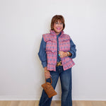 Woman wearing a pink plaid vest over a blue denim outfit, holding a brown handbag against a white wall.