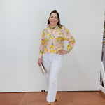 Woman wearing a floral blouse and white pants standing against a white wall.