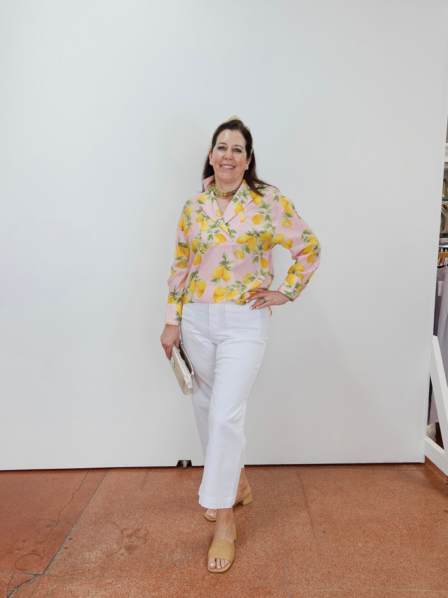 Woman wearing a floral blouse and white pants standing against a white wall.