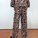 A person shown from the back wearing a matching leopard-print pajama set with a long-sleeve top and wide-leg pants, paired with black platform sandals, against a white wall and terracotta tiled floor.