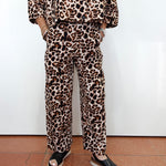 A person wearing a matching leopard-print lounge set with a cropped long-sleeve top and wide-leg pants, paired with black slide sandals, standing against a white wall.