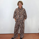 A woman with short blonde hair stands against a white wall wearing a matching two-piece leopard print pajama set with black slide sandals.