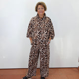 A woman with short blonde hair stands against a white wall wearing a matching two-piece leopard print pajama set with black slide sandals.