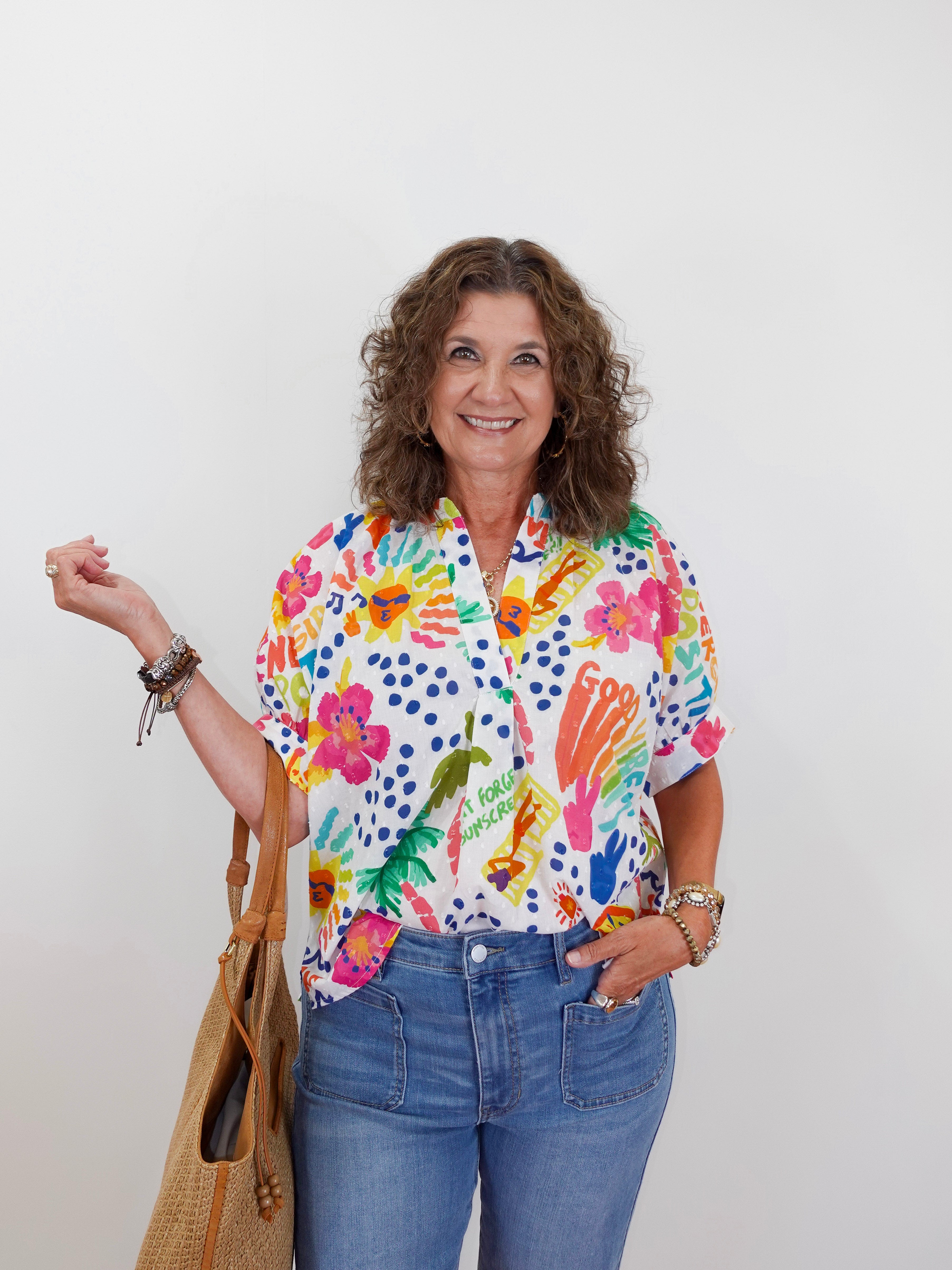 Woman wearing a colorful floral shirt with text, holding a straw bag against a white background