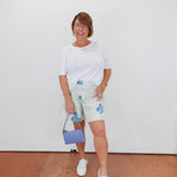Woman wearing a white top and floral shorts with a blue handbag against a white wall.