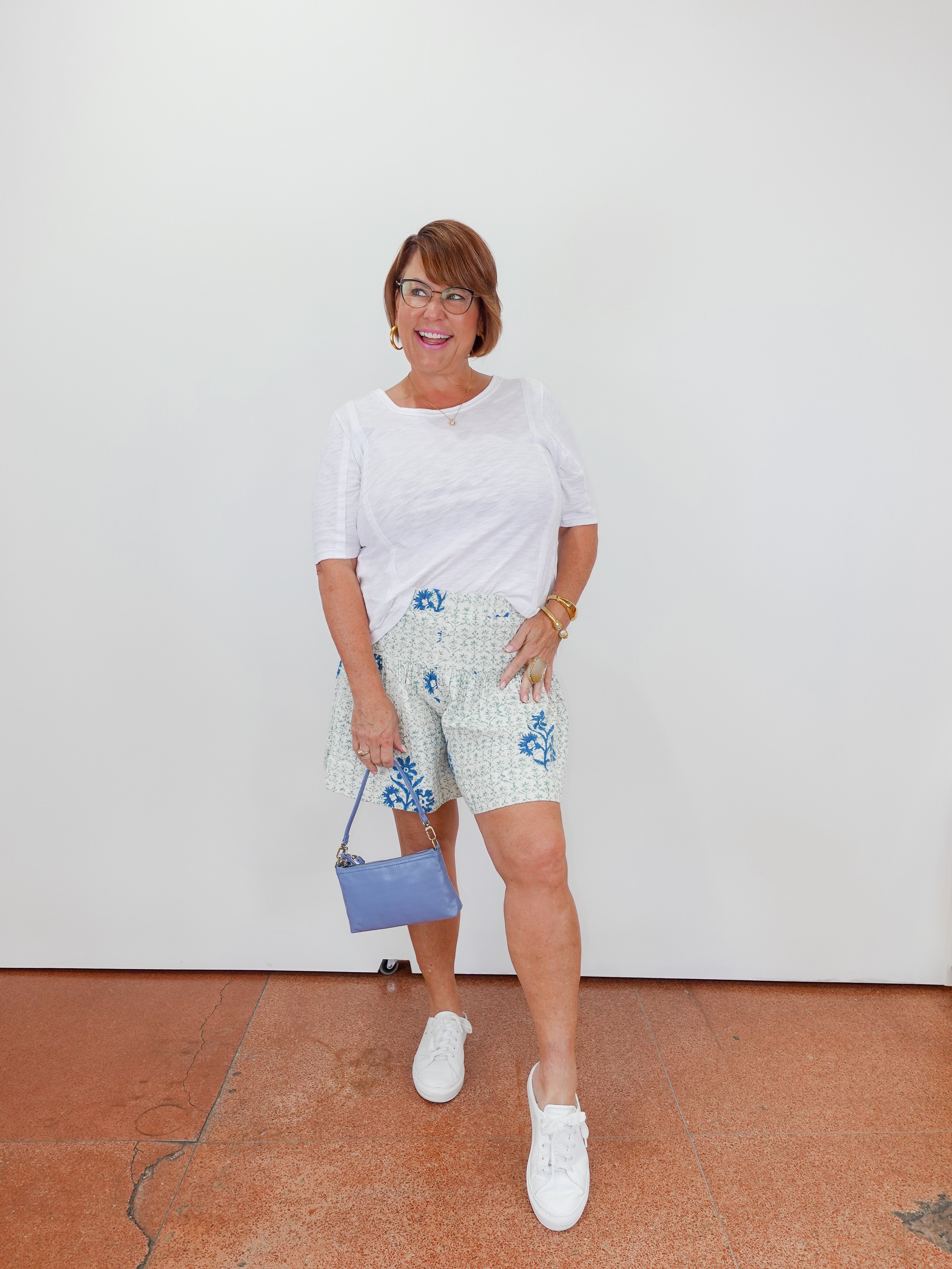 Woman wearing a white top and floral shorts with a blue handbag against a white wall.