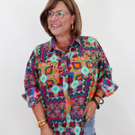 A woman wearing a colorful, patterned button-up shirt with geometric and bohemian designs, paired with light blue jeans, standing against a plain white background.