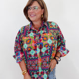 A woman wearing a colorful, patterned button-up shirt with geometric and bohemian designs, paired with light blue jeans, standing against a plain white background.