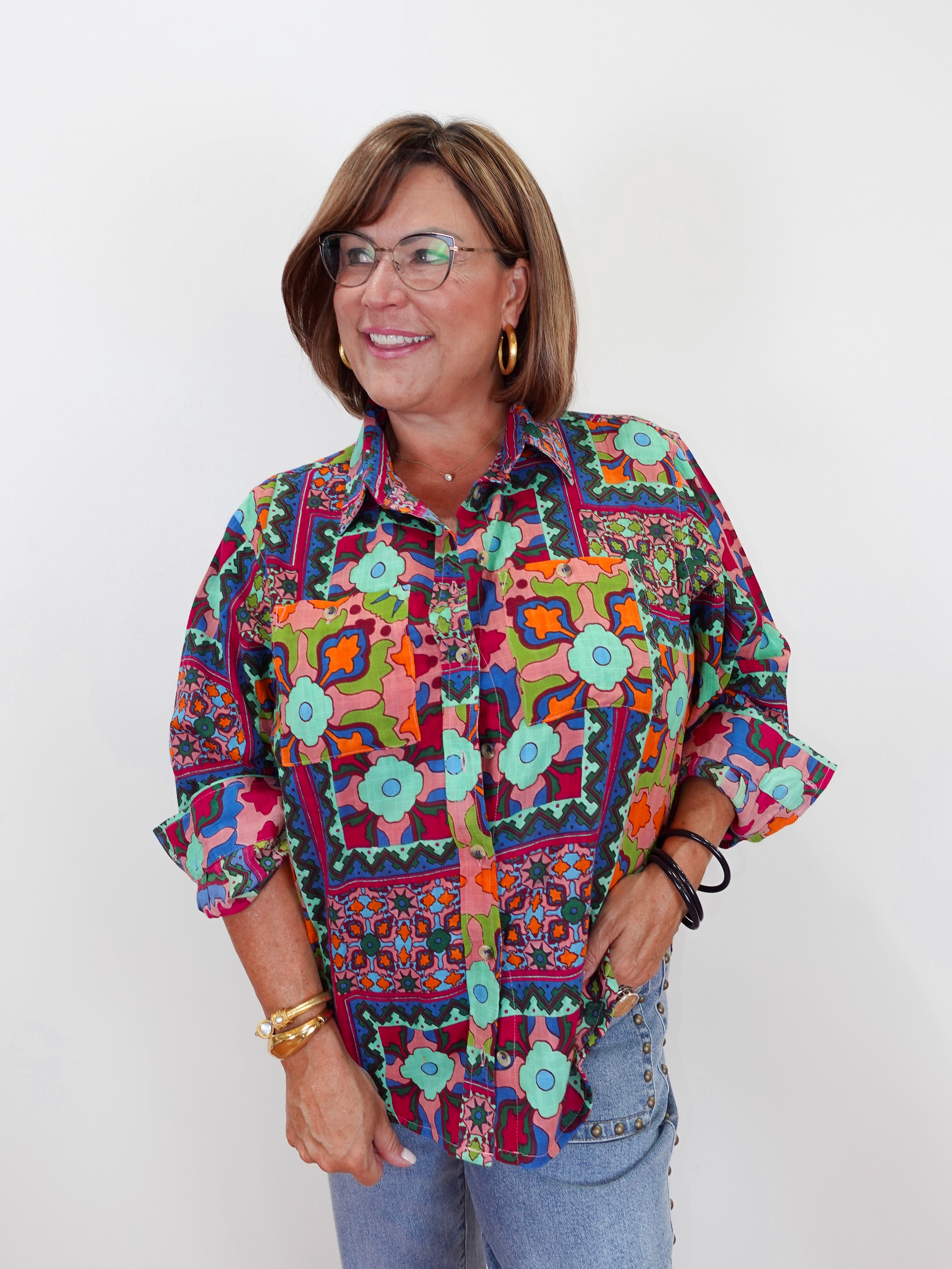 A woman wearing a colorful, patterned button-up shirt with geometric and bohemian designs, paired with light blue jeans, standing against a plain white background.