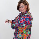 A woman with glasses and short brown hair models a colorful, long-sleeved button-up shirt with a vibrant geometric and floral pattern in pink, blue, green, orange, and yellow, paired with light blue jeans against a plain white background.