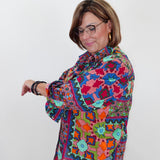 A woman with glasses and short brown hair models a colorful, long-sleeved button-up shirt with a vibrant geometric and floral pattern in pink, blue, green, orange, and yellow, paired with light blue jeans against a plain white background.