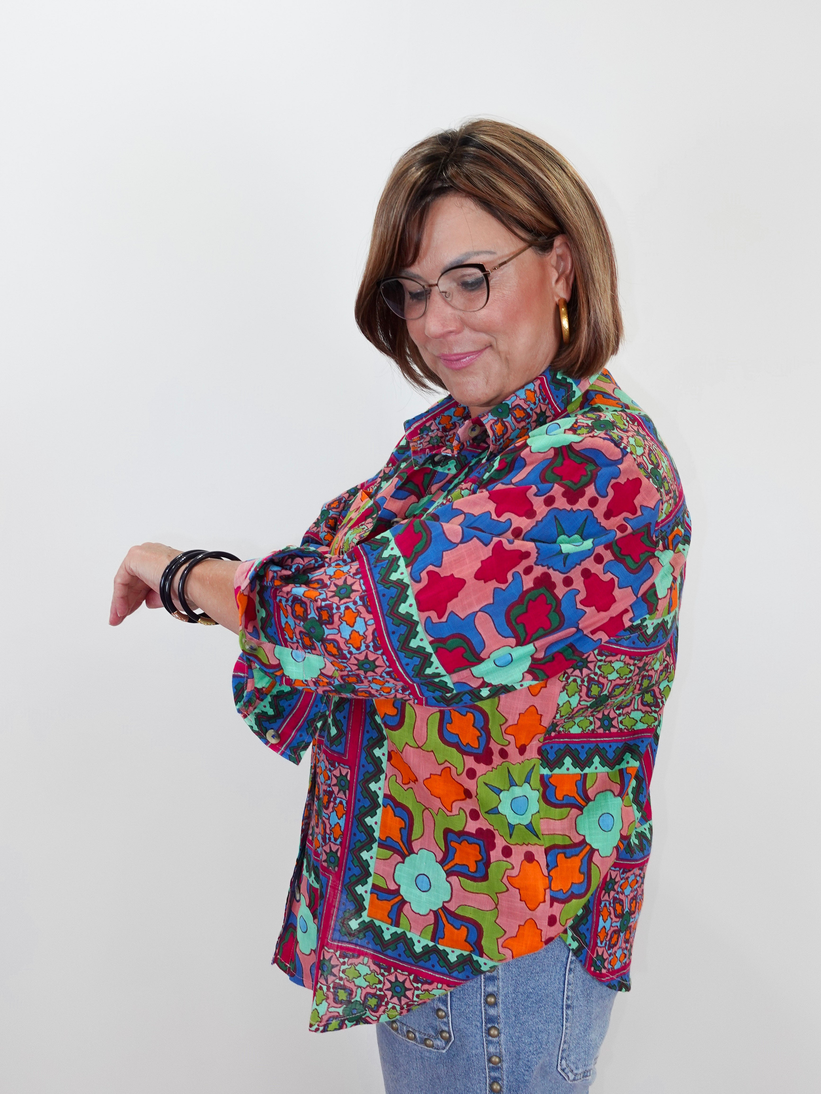A woman with glasses and short brown hair models a colorful, long-sleeved button-up shirt with a vibrant geometric and floral pattern in pink, blue, green, orange, and yellow, paired with light blue jeans against a plain white background.