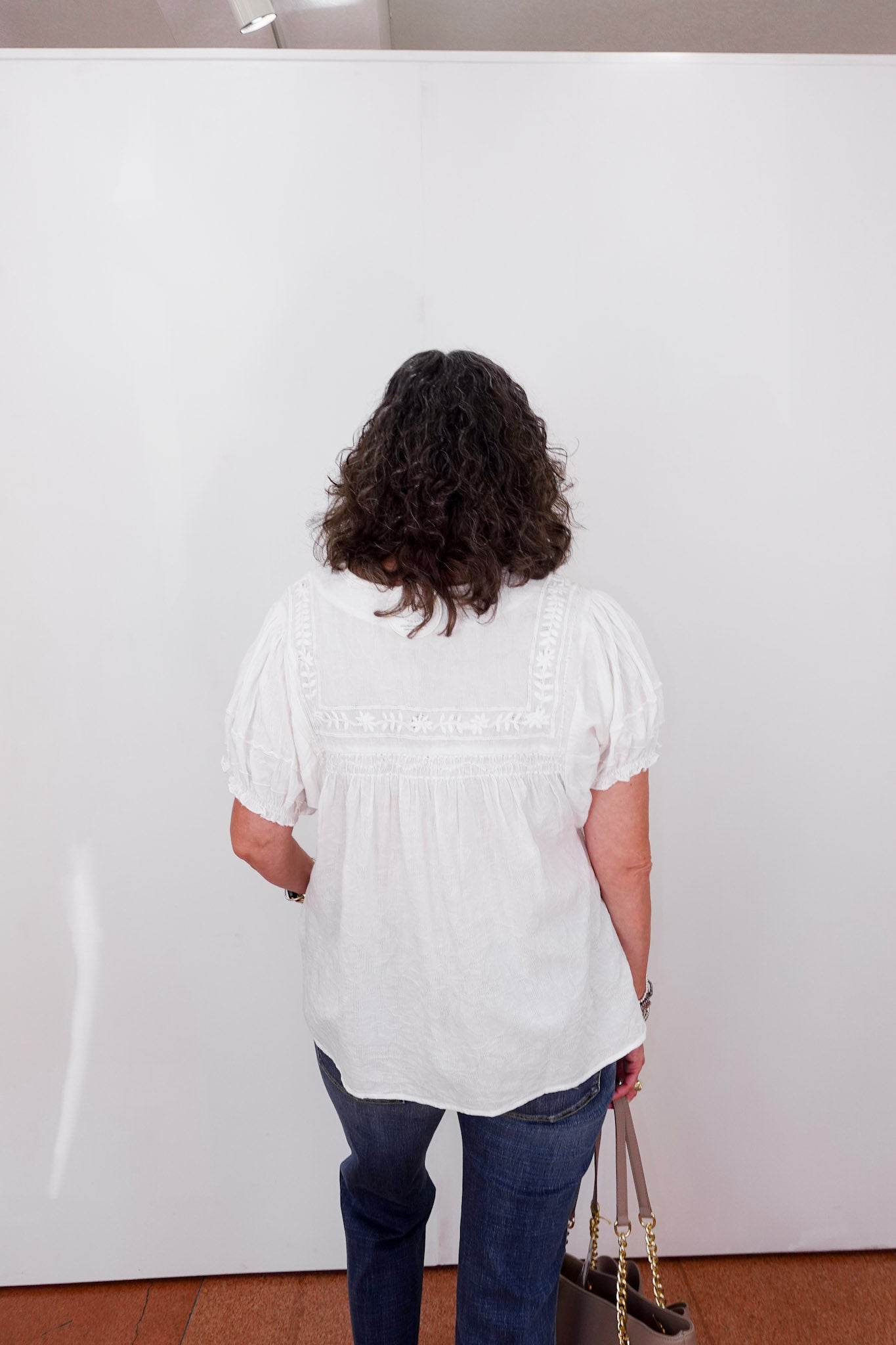 Person wearing a white blouse and blue jeans standing against a white wall.