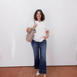 Woman wearing a white blouse, blue jeans, and holding a beige handbag against a white wall.
