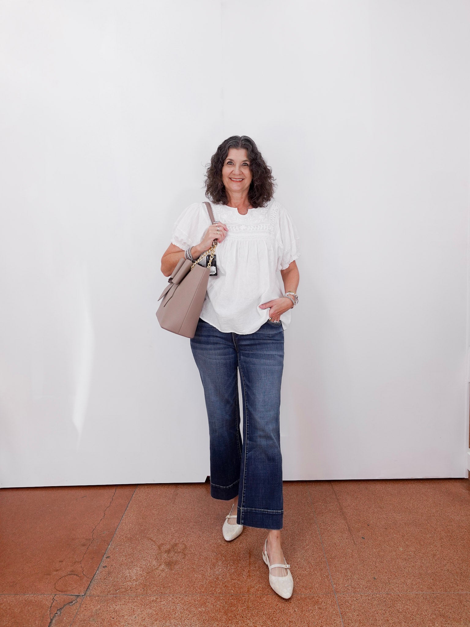 Woman wearing a white blouse, blue jeans, and holding a beige handbag against a white wall.